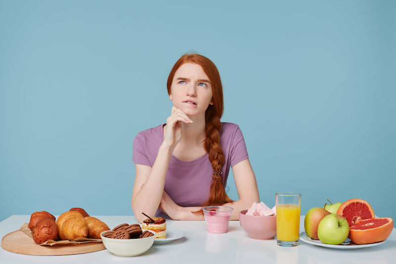 menina sentada em uma mesa com frutas doces e pães. Ela está com a mão no rosto e uma expressão de dúvida.