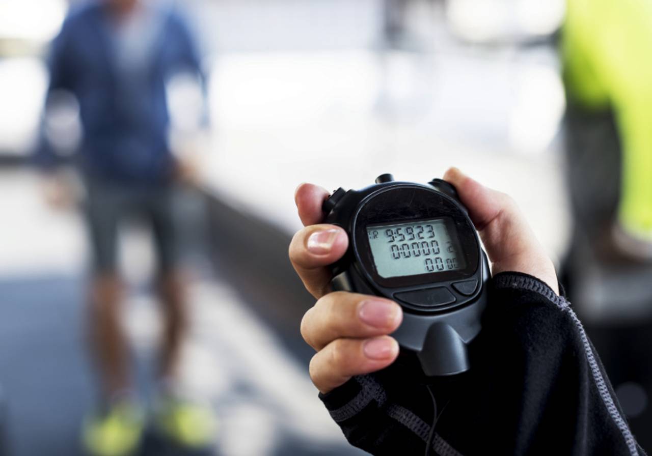 vista de frente aproximada de uma mão segurando um cronômetro enquanto calcula o pace de corredores ao fundo
