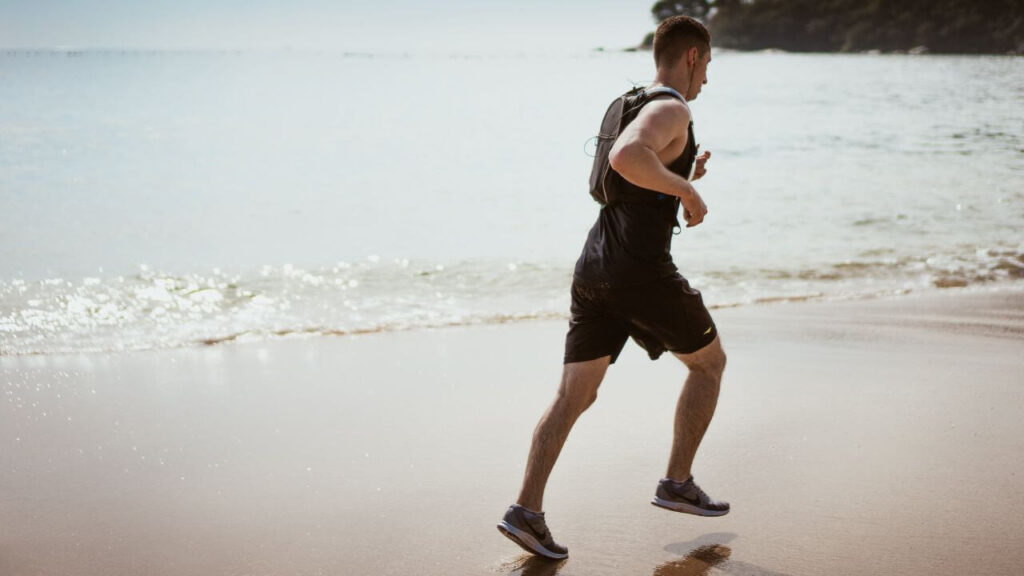 Homem correndo na areia da praia vestido de regata e short preto e com bolsa nas costas.