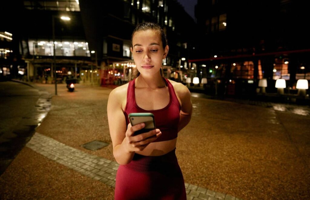 Mulher se preparando para correr, planejando rota em celular em área urbana e de noite